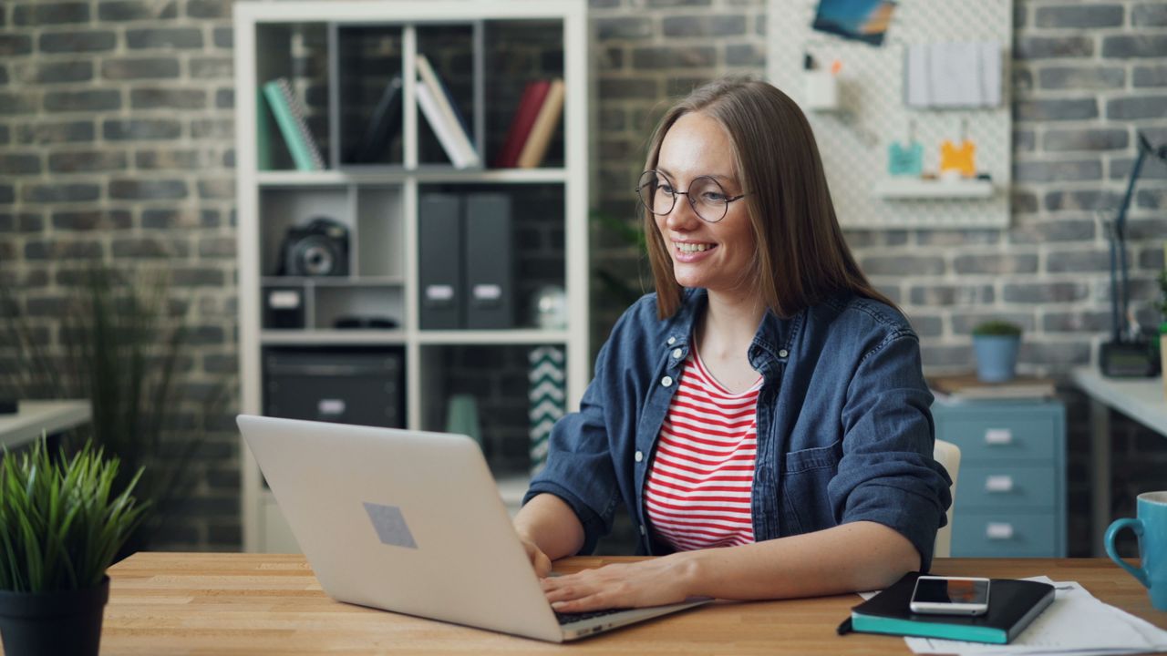 gallery-04 Cheerful woman working on laptop in modern office setting with smartphone and notebook.